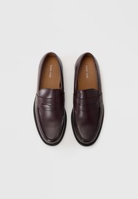 Mocassins en cuir bordeaux avec une texture lisse, bout rond, accent avec fente pour pièce de monnaie et semelle noire contrastante. Intérieur étiqueté "PIER ONE."