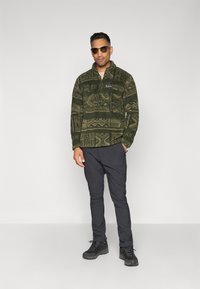 Man wearing patterned green fleece jacket, dark pants, black hiking shoes, and sunglasses standing against plain white background.