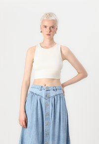 Lyhyeksi leikattu vaalean beige ribbineuletoppi, jossa on pyöreä pääntie, yhdistettynä korkeavyötarisään farkkuhameeseen, jossa on etupainikkeet ja flare-muotoinen design.