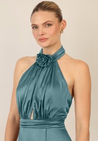 Abito halter in satin teal con un accento a forma di rosa sul collo, corpino plissettato e un'apertura in vita, caratterizzato da una superficie liscia e lucida.