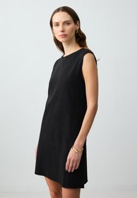Schwarzes ärmelloses Kleid mit Rundhalsausschnitt, taillierter Passform und glatter Textur, ausgestattet mit seitlichen Taschen und einem knielangen Saum. Goldene Armbänder setzen Akzente.