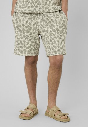 Homme portant un short en maille beige à motifs, les mains dans les poches et des sandales à brides beiges sur un fond gris clair.