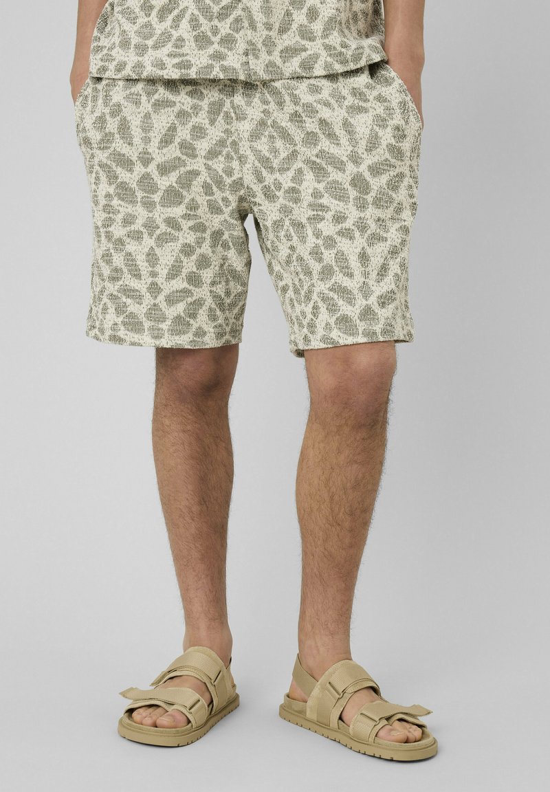 Homme portant un short en maille beige à motifs, les mains dans les poches et des sandales à brides beiges sur un fond gris clair.