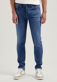 Moški modri jeans v ožjem kroju, z petimi žepi, diskretnim zbledeljem in kovinskimi_detajli. V kombinaciji z belimi in sivimi športnimi copati.