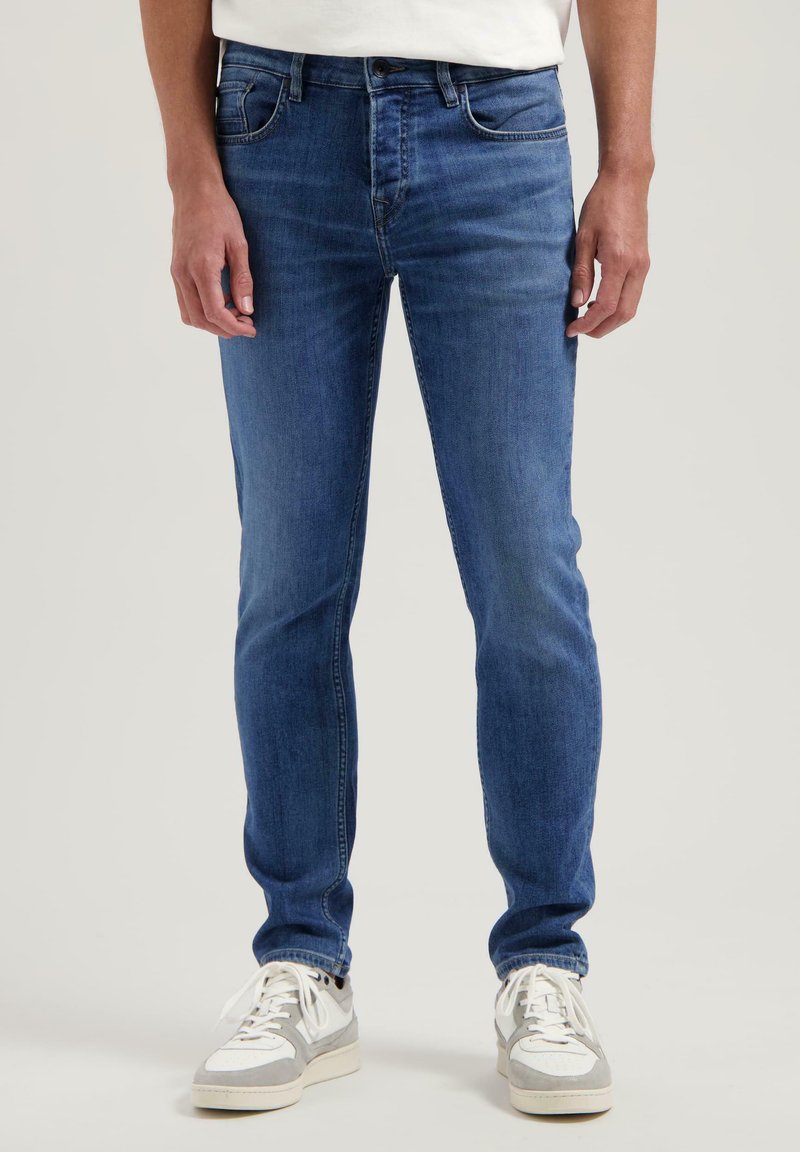 Moški modri jeans v ožjem kroju, z petimi žepi, diskretnim zbledeljem in kovinskimi_detajli. V kombinaciji z belimi in sivimi športnimi copati.