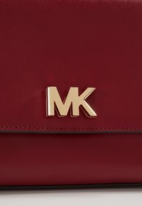 Burgundin värinen nahkalaukku, jossa on kultainen "MK" logo. Sileä pinta, tyylikäs muotoilu ja minimalistiset ompelutiedot.
