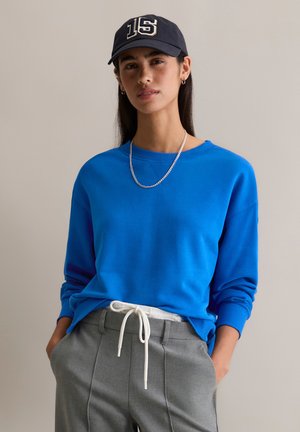 Blaues Sweatshirt mit gerippten Bündchen und Halsausschnitt, kombiniert mit grauen Hosen mit weißer Kordel. Eine dunkle Mütze mit der "15"-Detail.