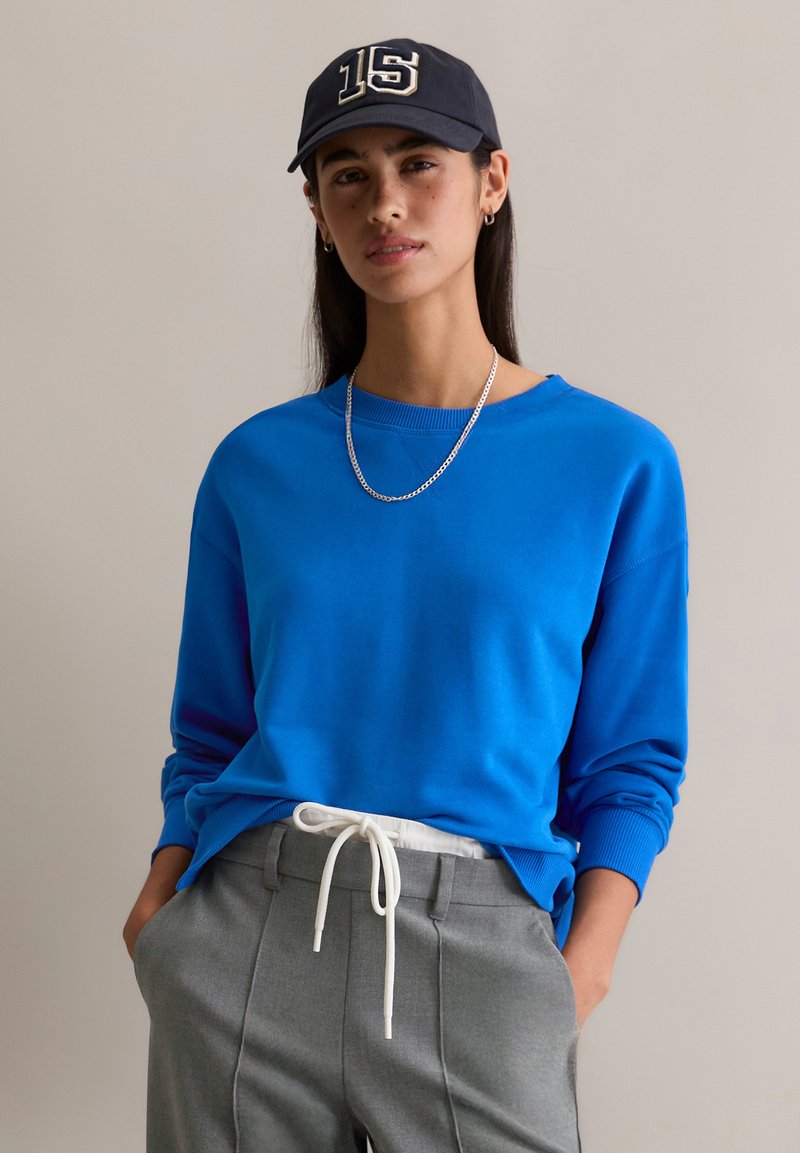 Niebieska bluza z prążkowanymi mankietami i dekoltem, zestawiona z szarymi spodniami z białym ściągaczem. Ciemna czapka z detalem "15".