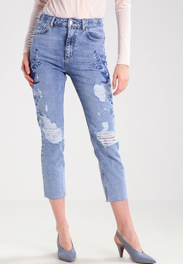 Světle modré džínové kalhoty s vysokým pasem, odřenými detaily, výšivkou na levé straně a zkrácenou délkou s třepeným lemem.