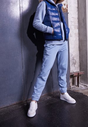 Persoon die lichtblauwe joggingbroek en hoodie draagt met een donkerblauw gewatteerd vest en witte sneakers, staand tegen een grijze industriële muur.