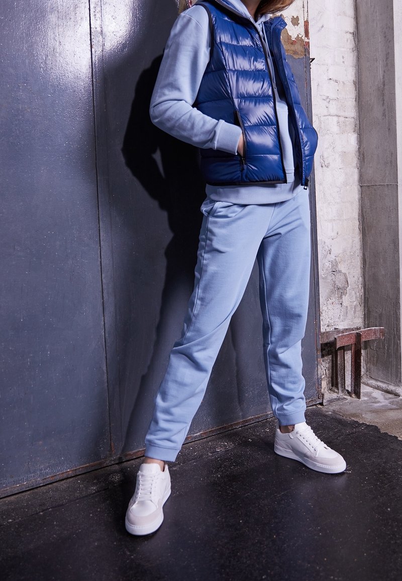 Personne portant un pantalon de survêtement bleu clair et un sweat à capuche avec un gilet matelassé bleu foncé et des baskets blanches, debout contre un mur industriel gris.