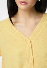 Kobieta w miękkim żółtym kardiganie z dekoltem w serek, z dwoma widocznymi guzikami, srebrną kolczykiem-kołem i prostymi ciemnymi włosami.