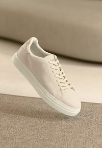 Beige mockasinsneakers med platta snören, rund tå och vit gummisula. Minimalistisk design, lätt silhuett och ingen synlig varumärkeslogotyp.