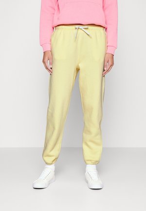 Personne portant un pantalon de jogging jaune clair à cordon, des baskets blanches et un sweat à capuche rose court, debout devant un fond uni.