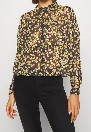 Vrouw draagt een transparante zwarte blouse met een kleine geelrode bloemenprint, gecombineerd met zwarte broek met hoge taille.