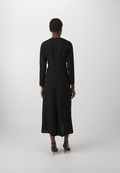 Vestido negro de manga larga con cuerpo ajustado y falda acampanada, que cuenta con una cremallera en la parte posterior. La tela tiene una textura suave. Se ven tacones negros.
