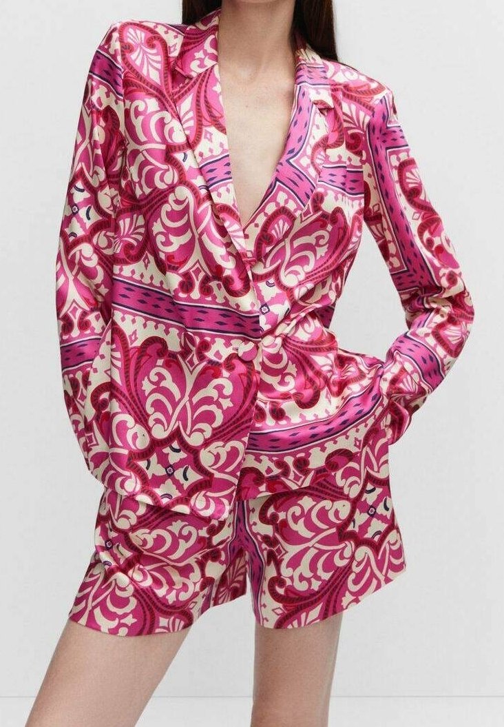 Femme portant un blazer rose et blanc à motifs avec un short assorti, posant avec une main dans la poche devant un fond uni.