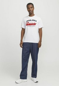 White cotton t-shirt with black and red text, including "JACK & JONES" and "Superior." Paired with loose, dark blue denim jeans and white sneakers.