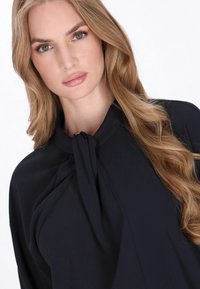 Blusa blu navy realizzata in un tessuto morbido, con un dettaglio al collo annodato e una vestibilità ampia. Le maniche lunghe e fluttuanti creano una silhouette rilassata.
