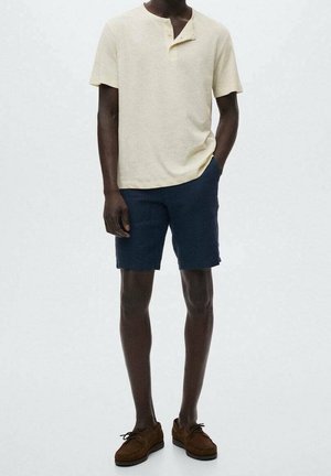 Jasno beżowa koszula henley z krótkim rękawem i trzema guzikami, w połączeniu z granatowymi szortami. Noszone są brązowe zamszowe buty. Minimalistyczny design.