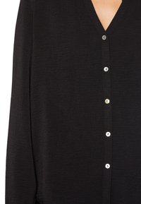 Camisa negra de manga larga con escote en V y cierres de botón plateados en el frente. El material es texturizado y ligeramente arrugado.