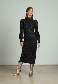 Vrouw die een zwarte lange mouwen jurk met pailletten en opgeblazen schouders draagt, gecombineerd met zilverkleurige metalen laarzen tot halverwege de kuit, staand op een betonnen vloer.