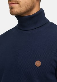Pull en col roulé bleu marine foncé avec texture côtelée et un patch en cuir marron rond sur la poitrine. Vue rapprochée du col.