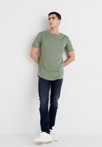 Camiseta verde de manga corta, con cuello redondo, combinada con jeans de color azul oscuro y zapatillas blancas, de pie contra un fondo blanco.