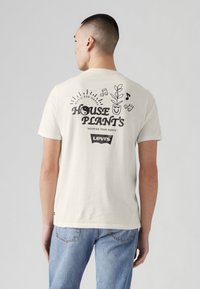 Tricou alb din bumbac, cu un design grafic de plante, textul „HOUSE PLANT'S” și note muzicale pe spate. Branding-ul de retail este vizibil.