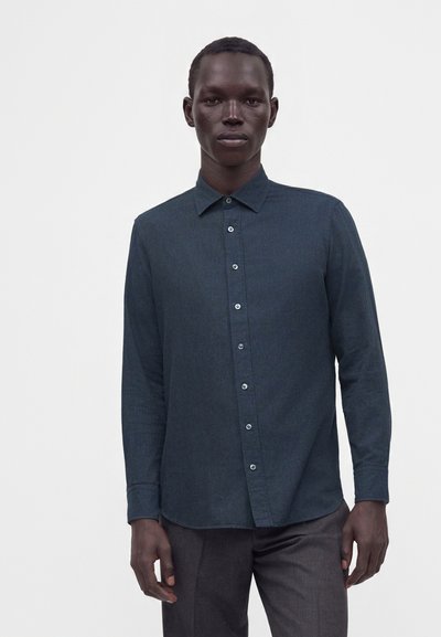 Homme portant une chemise boutonnée à manches longues bleu sarcelle foncé et un pantalon foncé, debout devant un fond blanc uni.