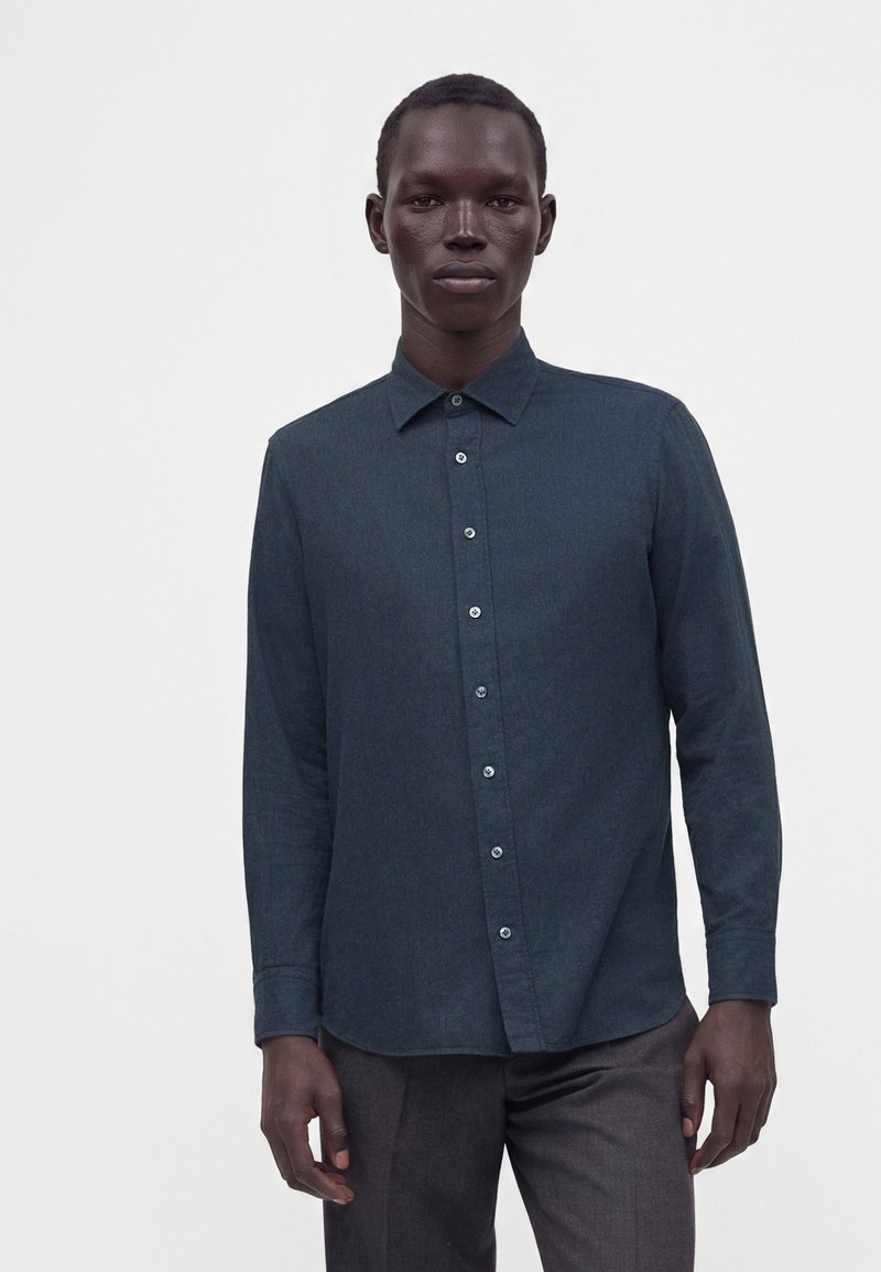 Homme portant une chemise boutonnée à manches longues bleu sarcelle foncé et un pantalon foncé, debout devant un fond blanc uni.