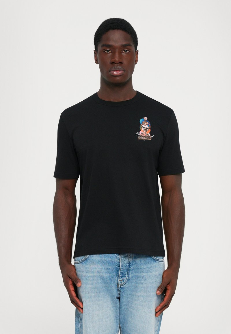 Man wearing black T-shirt with small colorful cobra design and text on upper left chest, paired with light blue jeans, standing straight.
