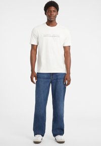 Balta trumpa rankovėmis marškinėliai su "GUESS JEANS" logotipu, derinami su laisvais mėlynais džinsais ir baltais sportbačiais su pilkais akcentais.