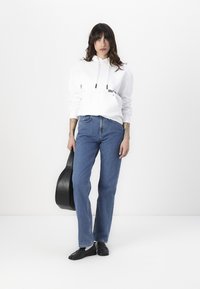 Sudadera blanca con cordones y logo, combinada con jeans azul de pierna recta. El modelo sostiene una guitarra negra, vistiendo zapatos negros.