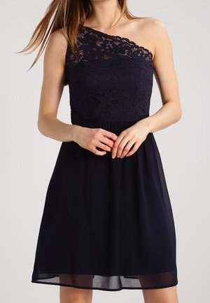 Femme portant une robe bleu marine à une épaule avec un corsage en dentelle florale et une jupe fluide, debout devant un fond clair uni.