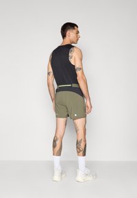 Svart ermeløs topp, olivengrønne shorts med to hvite detaljer og et grønt belte. Hvite sokker og lyse joggesko fullfører antrekket.