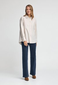 Jasno beżowa koszula na guziki z kołnierzykiem, długimi rękawami i zaokrąglonym dołem, zestawiona z ciemnoniebieskimi spodniami dzwonami i brązowymi botkami za kostkę.