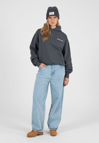 Hall halliga kapuutsiga dressipluus eest taskuga, mida kannavad helesinised laia säärega teksad ja pruunid jalanõud. Modellil on peas soonikkoeline hall müts.