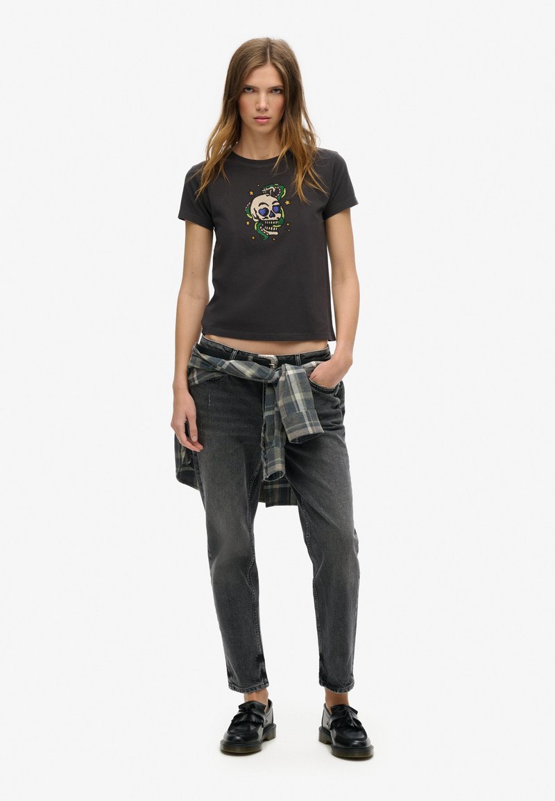 Czarna krótka koszulka z kolorowym haftem czaszki, w połączeniu z szarymi dżinsami. Dodatki to koszula w kratę związana w pasie. Czarne buty.