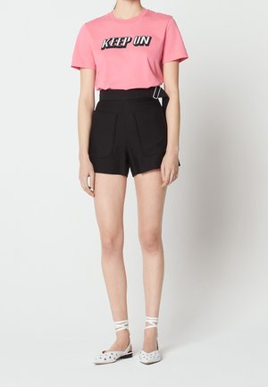 Person iført en lyserød t-shirt med teksten "KEEP ON", højttaljede sorte shorts med forlommer og hvide flade sko med ankelstropper.