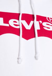 Felpa bianca con cappuccio caratterizzata da un prominente logo rosso di Levi's, con testo bianco e coulisse. Il materiale è in cotone morbido, la texture appare liscia.