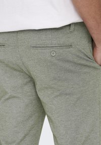 Pantalones de color verde claro con textura, de corte entallado, y un bolsillo trasero con un botón a juego.