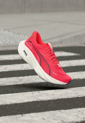 Chaussure de course Puma rouge vif avec semelle blanche en l'air au-dessus d'un passage piéton sur une surface de rue.