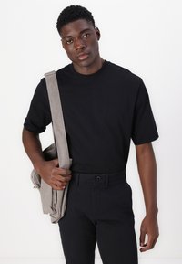 Black short-sleeve t-shirt paired with dark pants, holding a light gray canvas messenger bag with adjustable strap and button details.