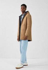 Chaqueta acolchada marrón con capucha, acolchado vertical y forro negro, combinada con jeans azul claro y zapatillas blancas. Diseño minimalista.