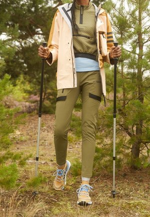 Osoba ubrana w oliwkowe spodnie i kurtkę turystyczną, beżowy płaszcz przeciwdeszczowy oraz buty trekkingowe, używająca kijków trekkingowych w lesie.