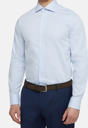 Mann in hellblauem, taillierten Hemd, das in eine marineblaue Hose gesteckt ist, mit einem dunkelbraunen Gürtel, der vor einem einfarbigen Hintergrund steht.
