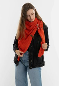 Rode gebreide sjaal met een gemêleerd patroon, voorzien van een houten sluiting en een verstelbare band, gedragen met een zwarte jas en blauwe jeans.