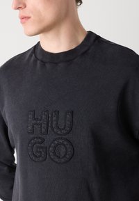 Czarny sweter z ribbedowym dekoltem i teksturowanym logo "HUGO" w wytłoczonym wzorze. Gładki materiał z długimi rękawami.