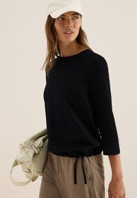 Femme portant une casquette blanche, un haut noir à manches 3/4 avec un ourlet à cordon, un pantalon beige et tenant un sac en toile beige, regardant légèrement sur sa droite.